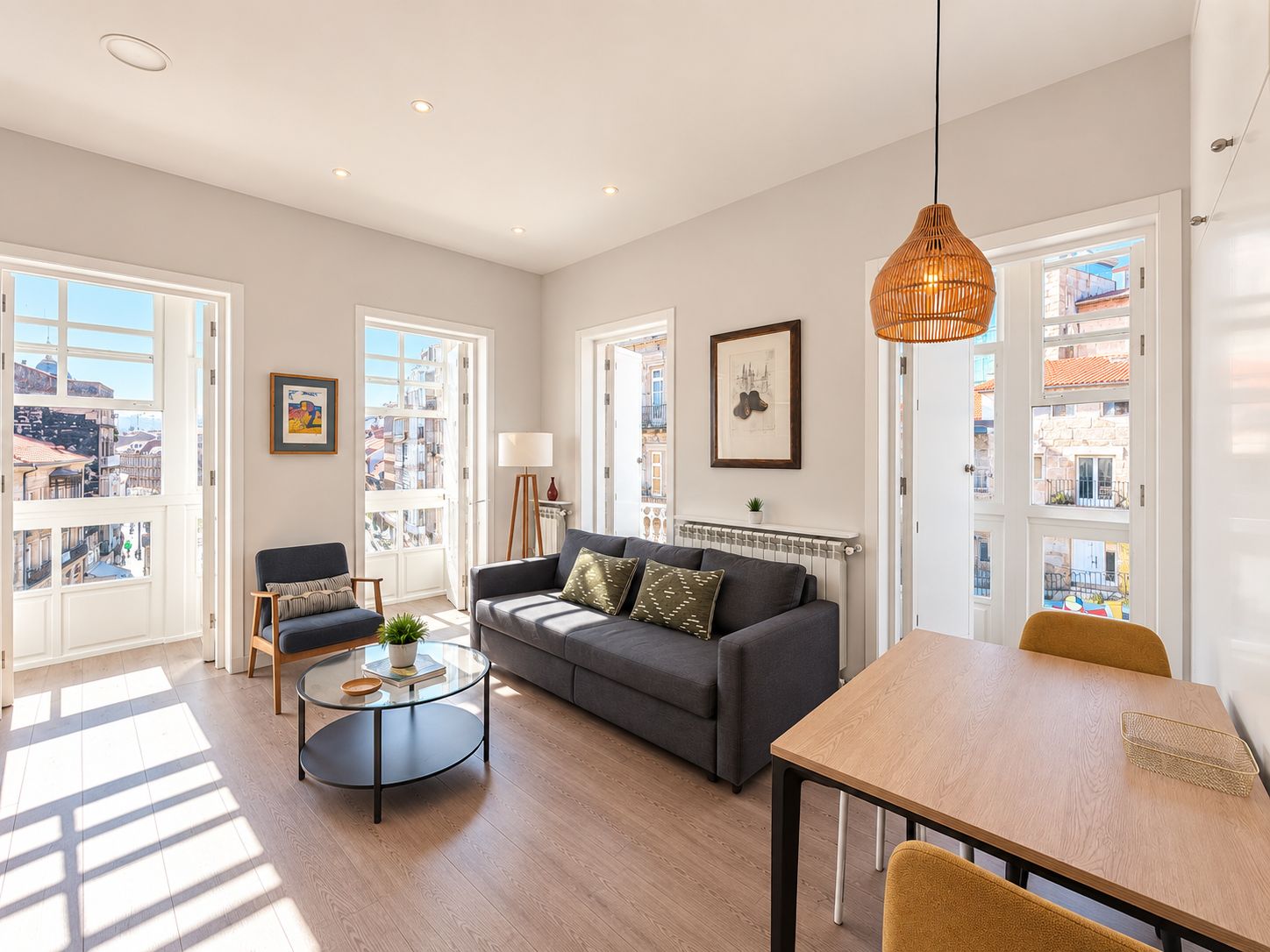 A spacious living room with a sofa, a coffee table, and a dining table. There are large windows providing a view of the city.