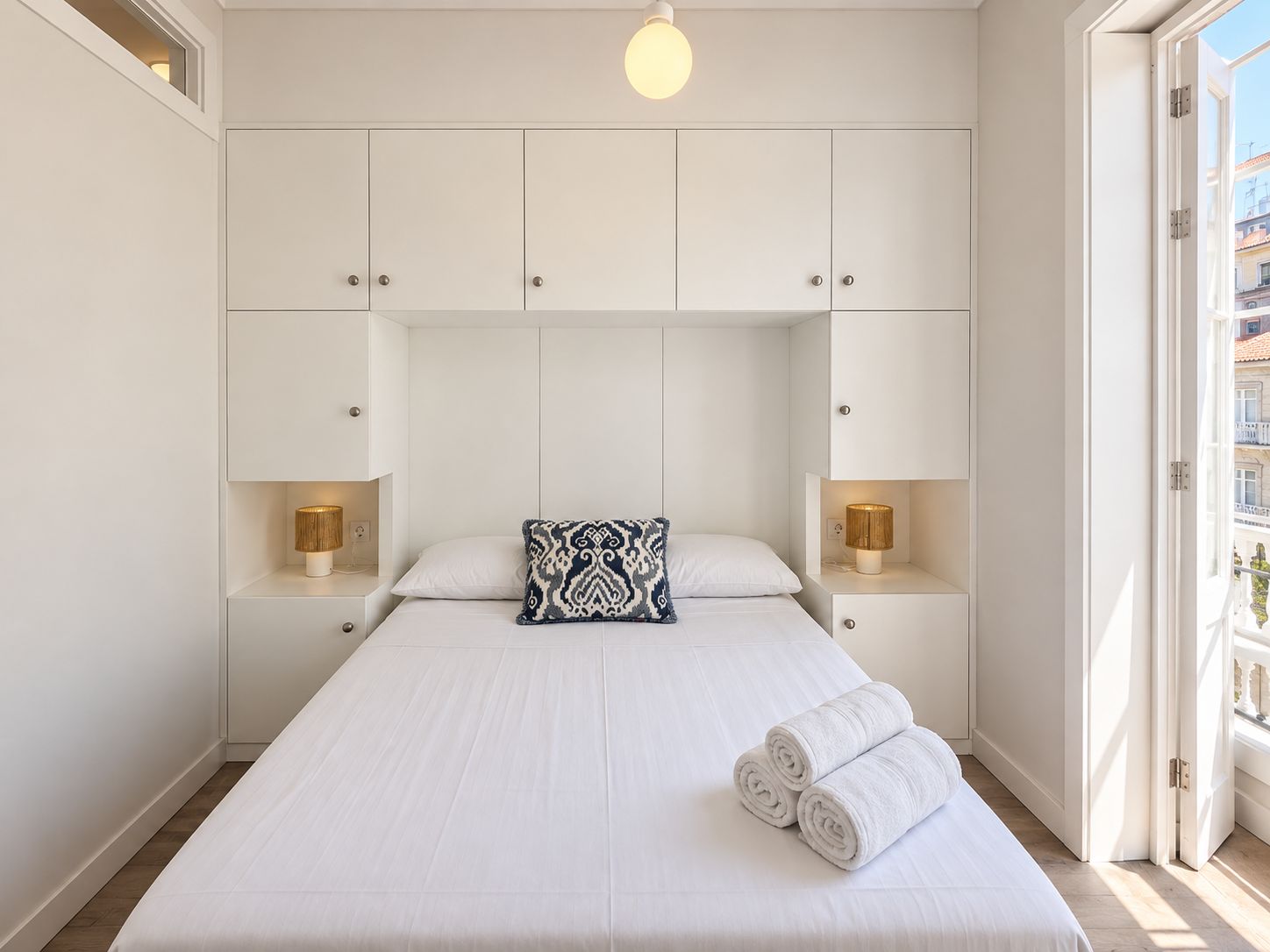 Una camera d'albergo con un letto grande, due lampade sui comodini, un cuscino decorativo e vista attraverso una porta aperta.