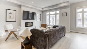 Uma sala de estar moderna com um sofá cinzento, uma poltrona branca, uma televisão de tela plana na parede e uma janela com vista para a cidade.