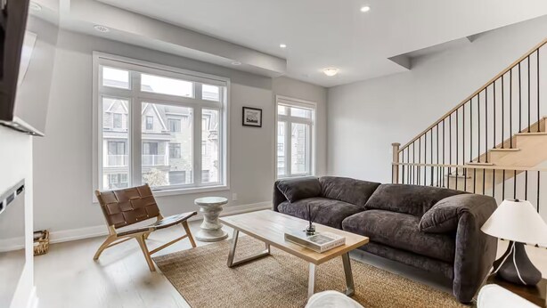 Uma sala de estar moderna com sofá, mesa de centro e uma poltrona. Há uma escada com corrimão e um abajur ao lado.