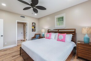 A bedroom with a large bed, a ceiling fan, a nightstand with a lamp, and a window with blinds. - Pili Mai 15F - newly remodeled (Poipu)