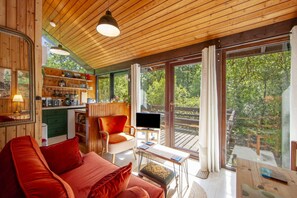 En ombonad inredning med ett träpanelat tak, en röd soffa, en tv och en balkong med utsikt över träd.