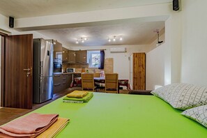 Una habitación compacta con colcha verde, un armario de madera, una zona de cocina con refrigerador, horno y microondas, y una mesa de comedor con sillas.