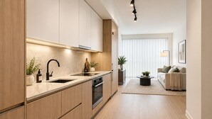A modern kitchen with wooden cabinets, a built-in oven, and a sink. There is a dining area with a sofa and a coffee table. - Toren Hotel & Suites (Toronto)