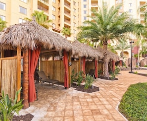 A resort with thatched-roof cabanas, lounge chairs, and a paved walkway. - Orlando | Pool & Disney Access | 2BR Suite (Lake Buena Vista)