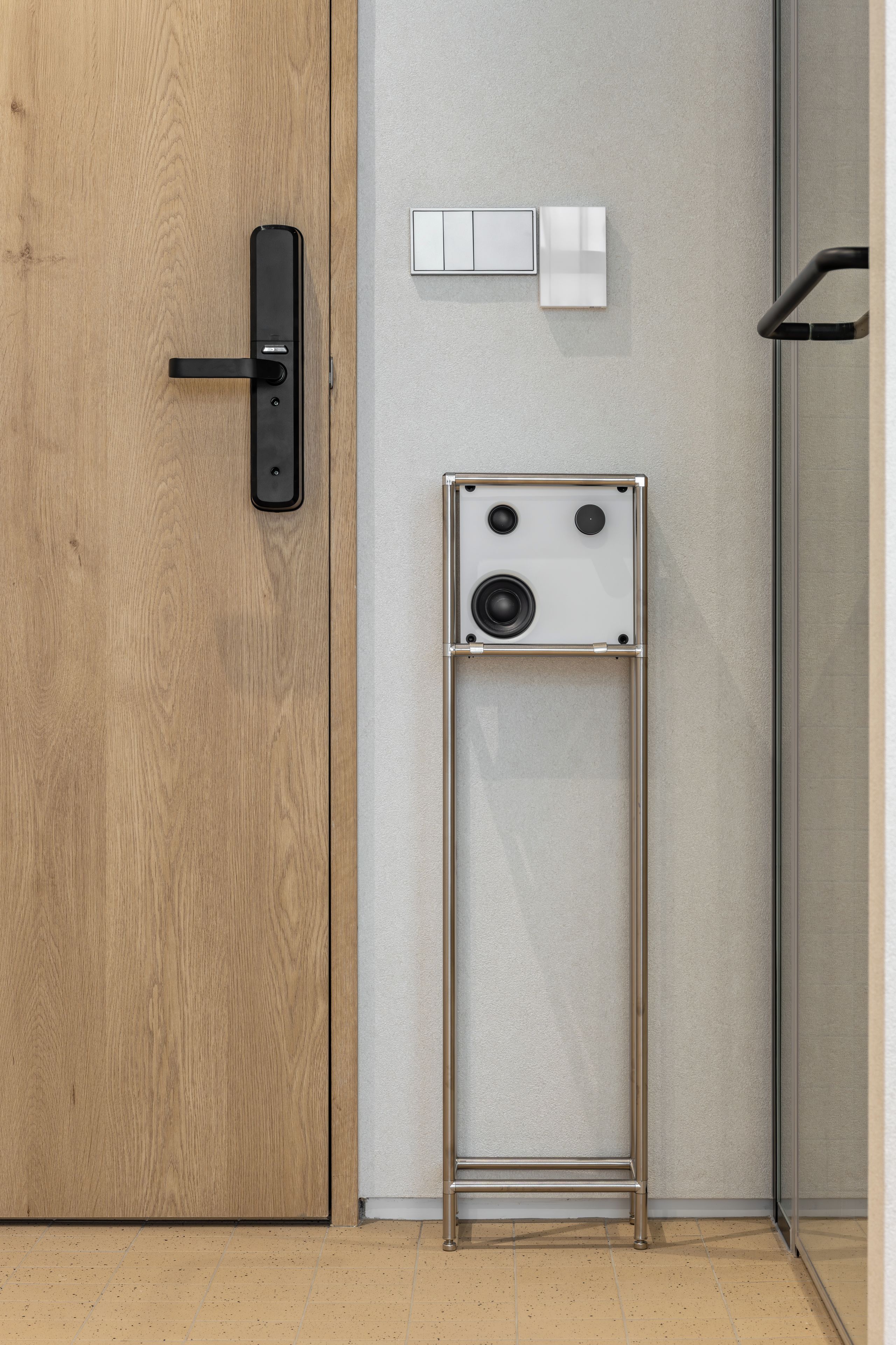 A wooden door with a black handle, a white wall with a thermostat, and a metal stand with a speaker.