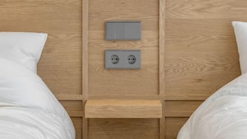 Een hotelkamer met een houten hoofdeinde, twee bedden met witte beddengoed, een klein nachtkastje en een wandcontactdoos.