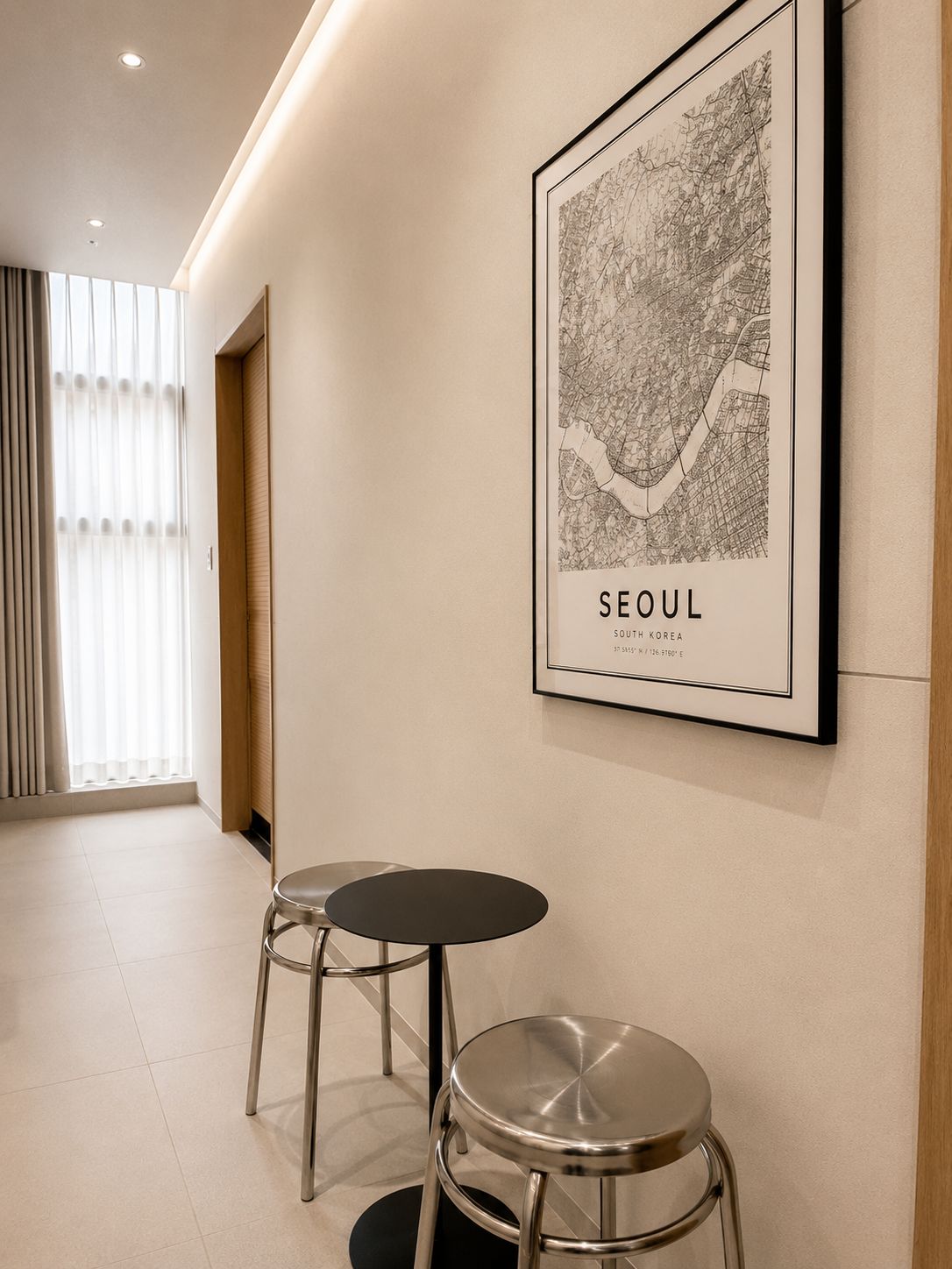 A hallway with a framed map of Seoul, two metal stools, and a door with blinds.