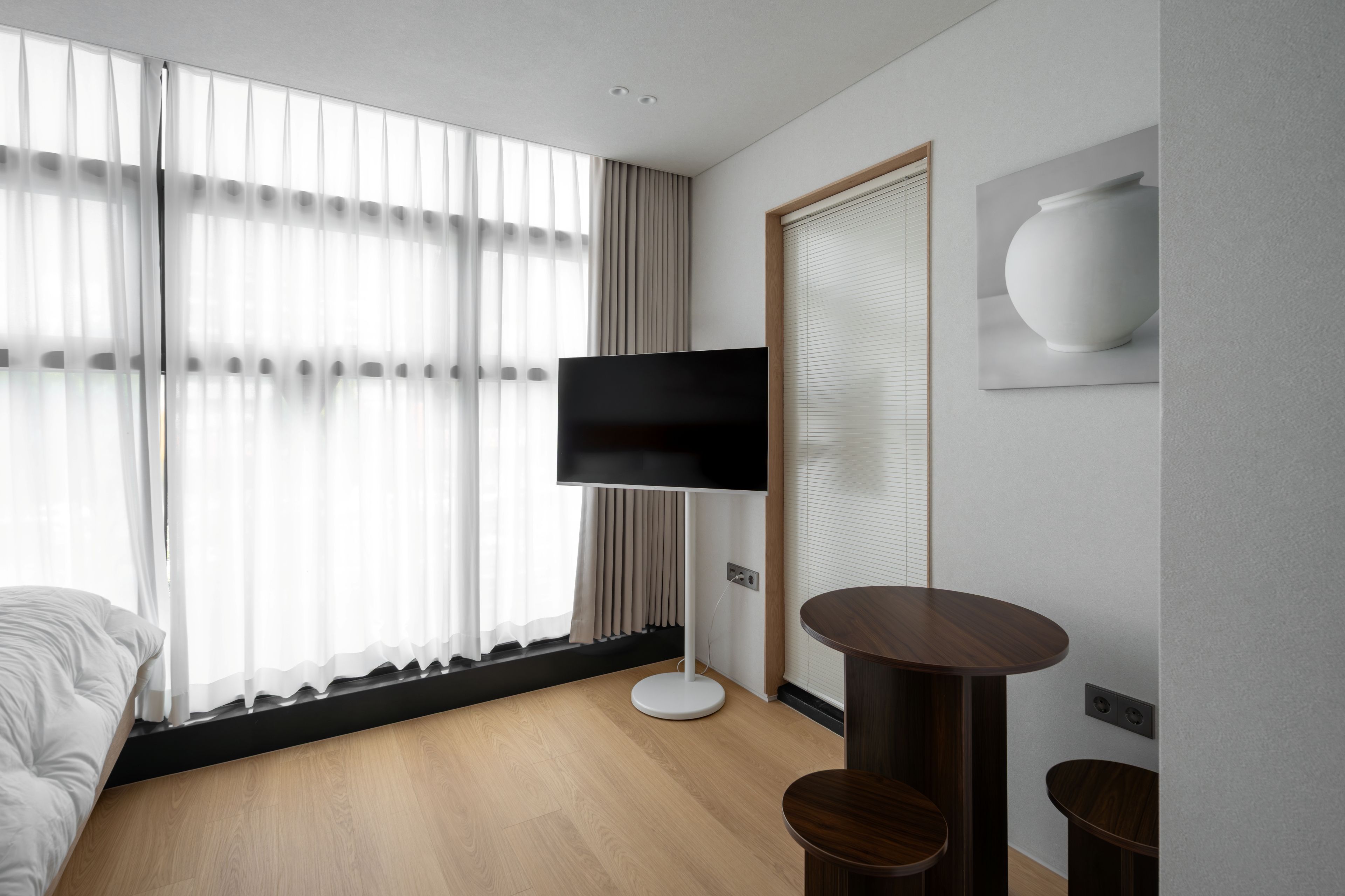 A modern hotel room with a bed, a flat-screen TV mounted on a stand, a round wooden table, and a large window with sheer curtains.