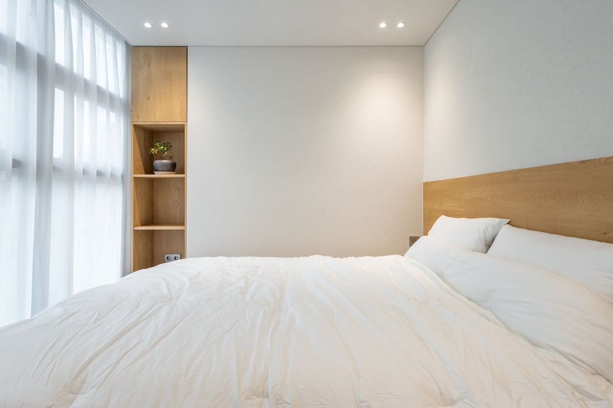 Une chambre avec un grand lit, une tête de lit en bois et des étagères intégrées, agrémentée d’une plante en pot.