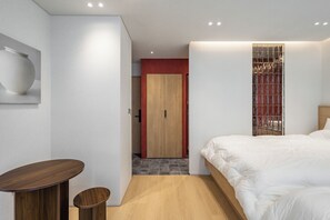 A modern bedroom with a bed, a wooden side table, a small stool, and a door leading to another room.
