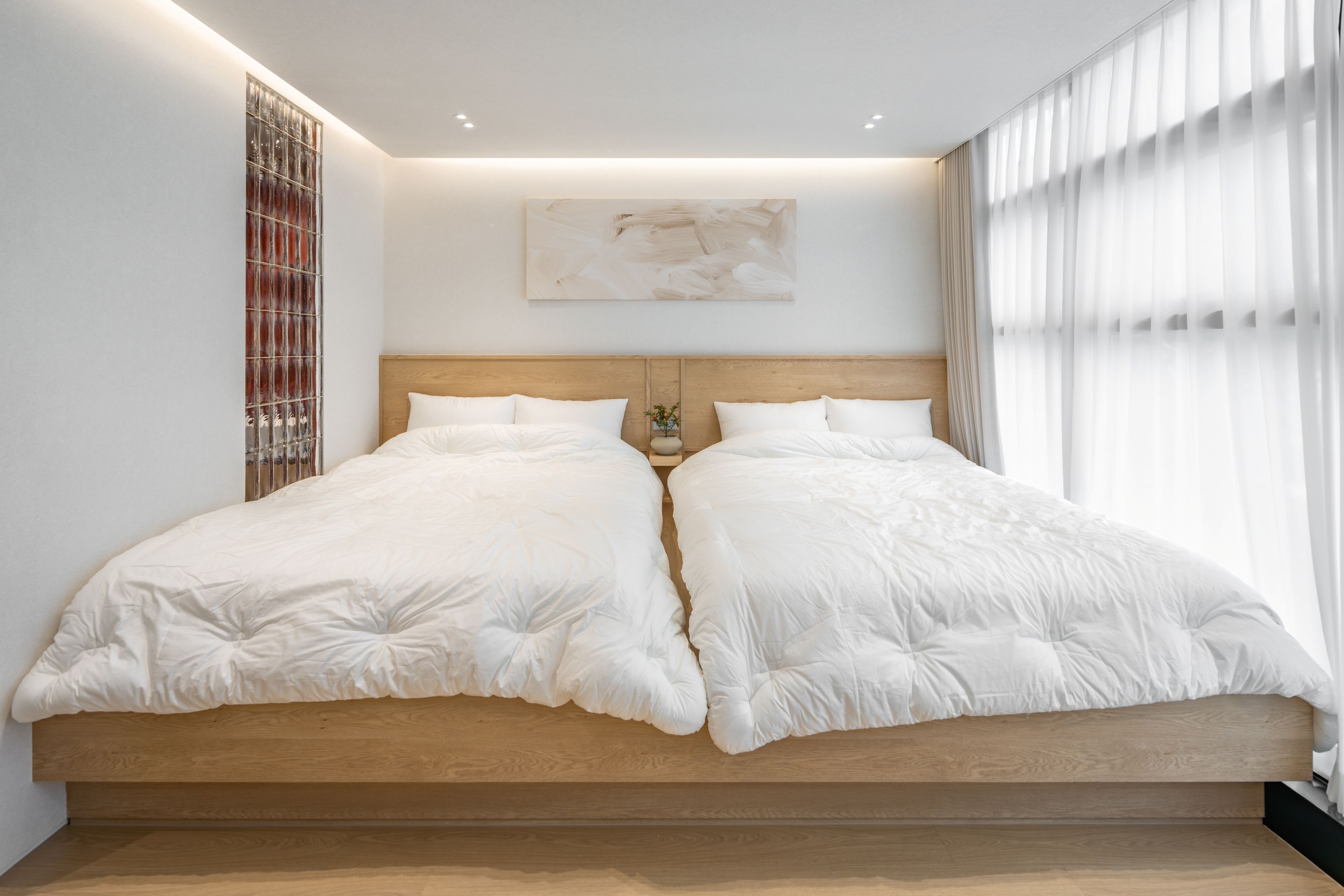 A double bed with a wooden headboard, a wall-mounted abstract painting, and a window with sheer curtains.