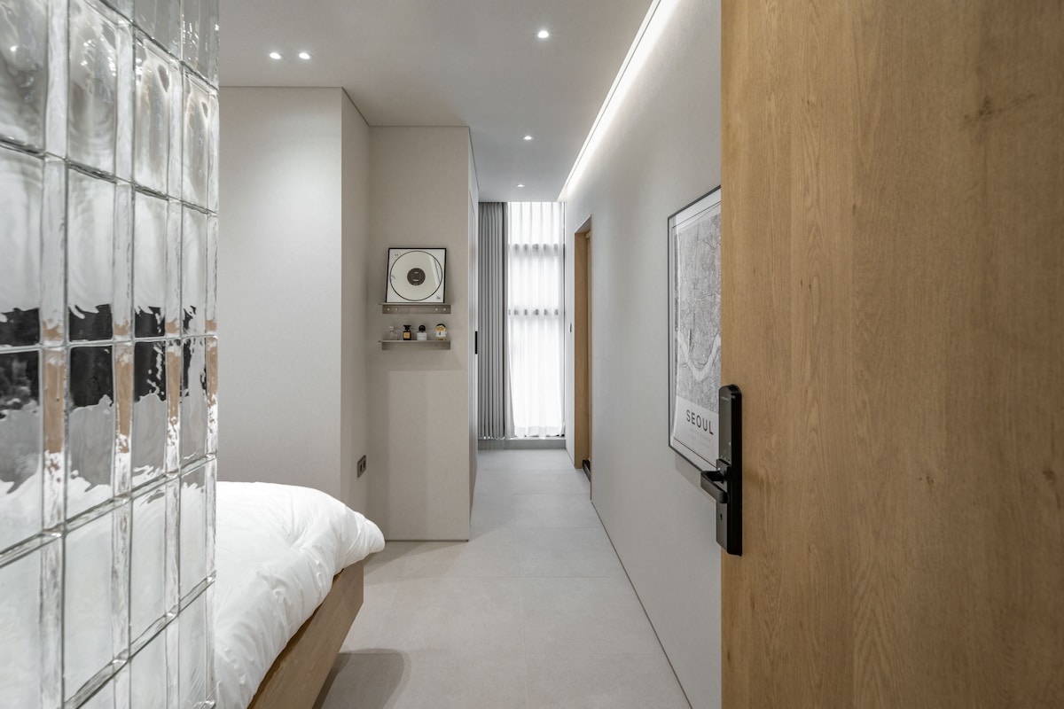 Une chambre d’hôtel moderne avec une cloison en verre, un lit, une porte en bois et un tableau encadré au mur.