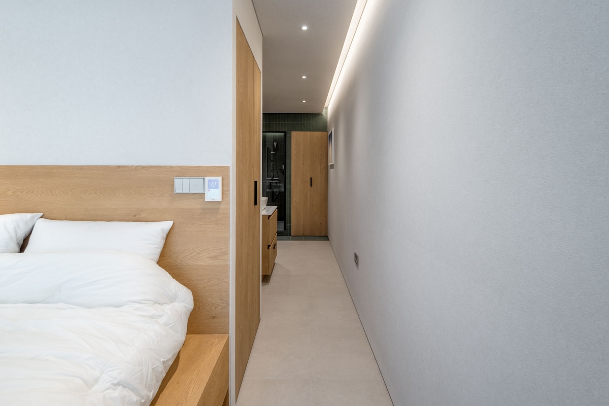 Habitació d'hotel amb un llit, un panell de fusta al capçal, un bany petit amb dutxa i un passadís que condueix a una altra habitació.
