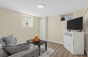 A modern living room with a sofa, a coffee table with a vase of flowers, a flat-screen TV on a white cabinet, and a window with blinds. - DC Area Apt w Fenced Yard - 1 Mi to Metro! (Silver Spring)