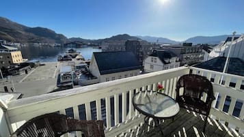 A balcony with wicker furniture, overlooking a harbor with buildings and mountains in the distance.