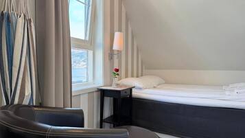 A hotel room with a bed, a chair, a bedside table with a lamp, and a window with curtains.