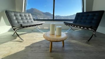 Habitación con dos sillones de cuero negro orientados hacia un ventanal, una pequeña mesa de madera con dos velas, y vistas a montañas.