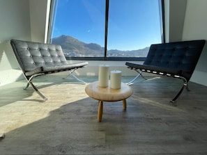 Habitación con dos sillones de cuero negro orientados hacia un ventanal, una pequeña mesa de madera con dos velas, y vistas a montañas.