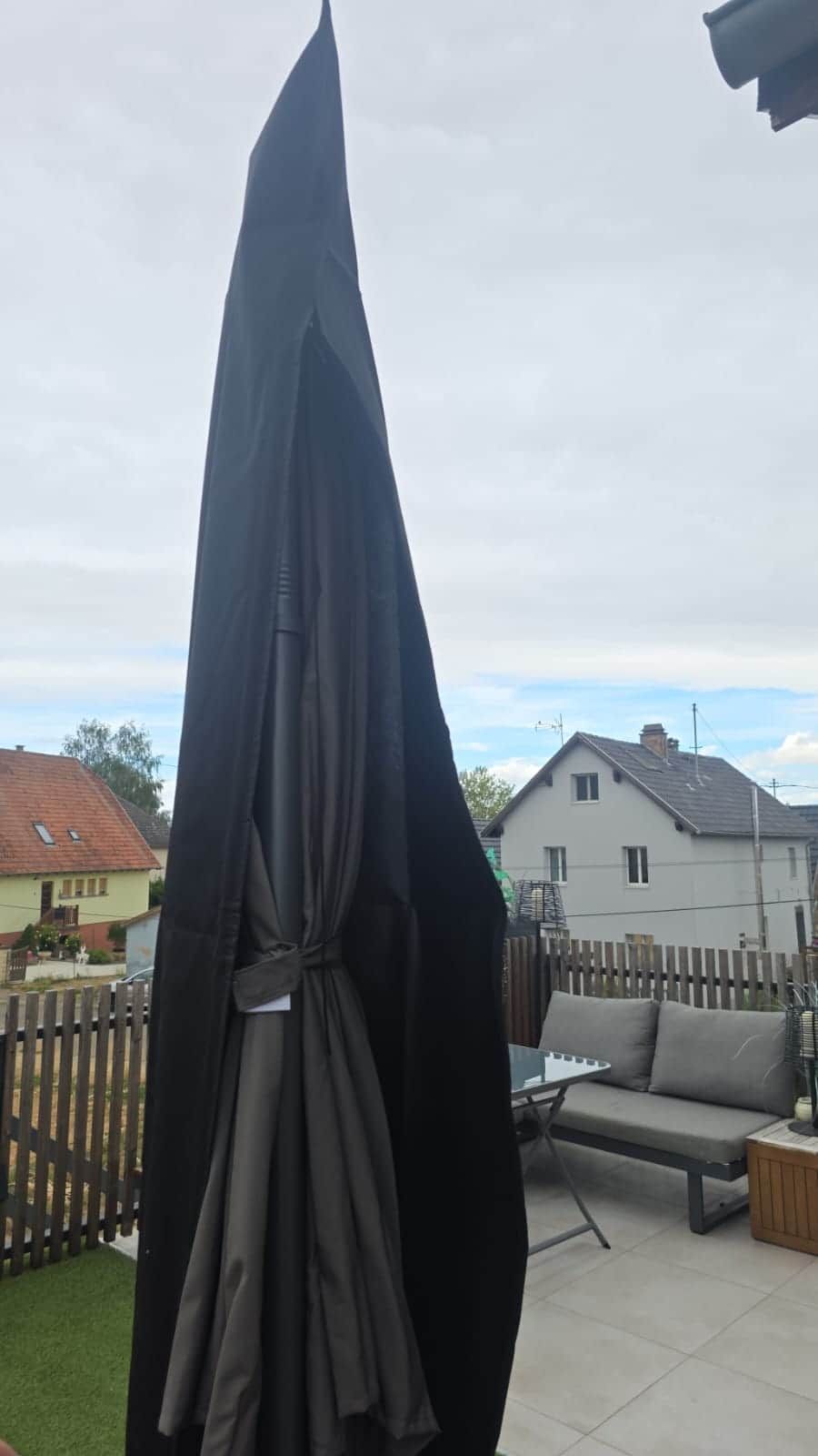 En udendørs terrasse med en sort parasol, et træhegn og et hvidt hus i baggrunden.