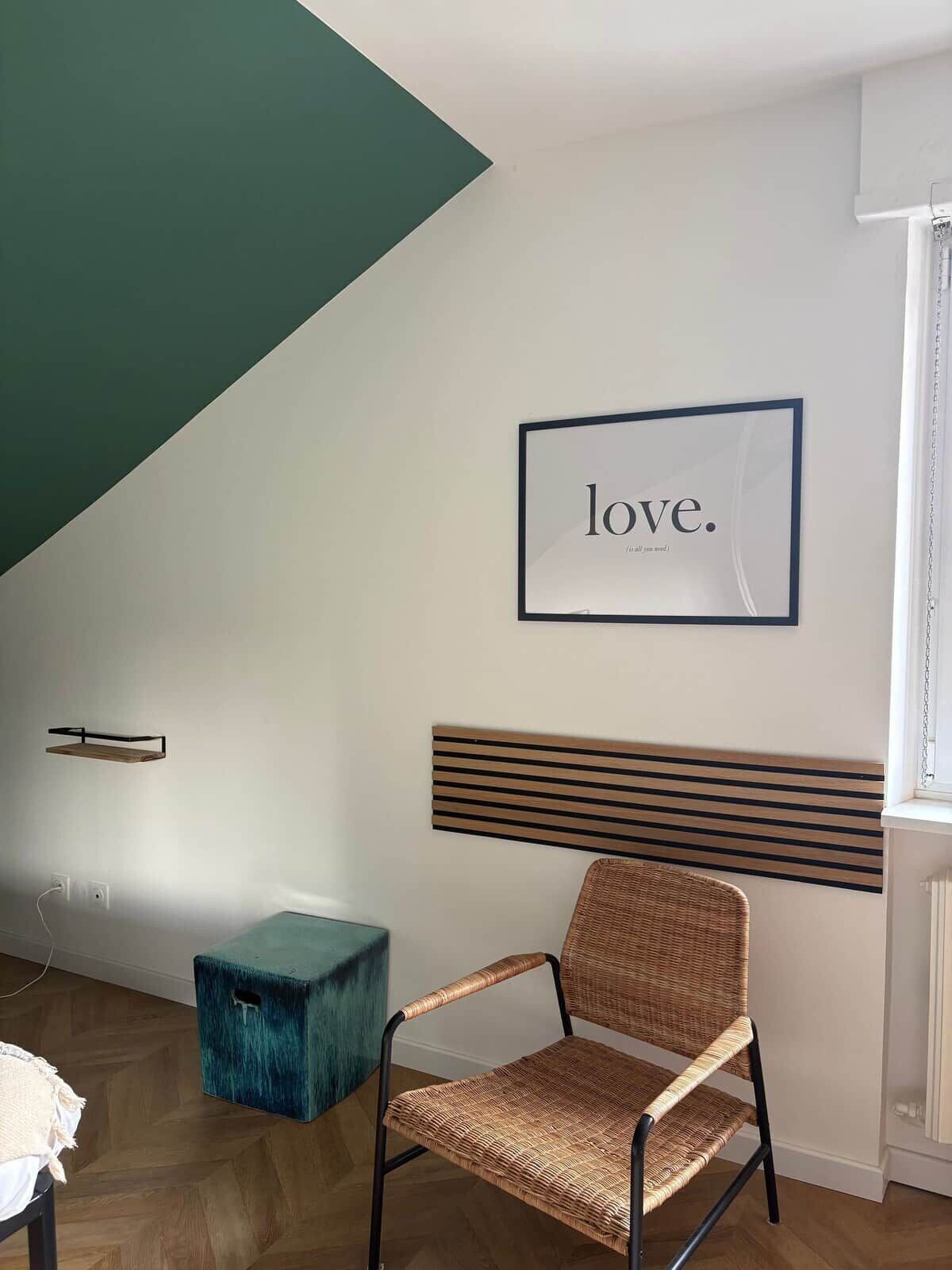 Une pièce avec un plancher en bois, une chaise en osier, une boîte de rangement de couleur turquoise, une étagère fixée au mur et un panneau encadré sur lequel on peut lire le mot « amour ».