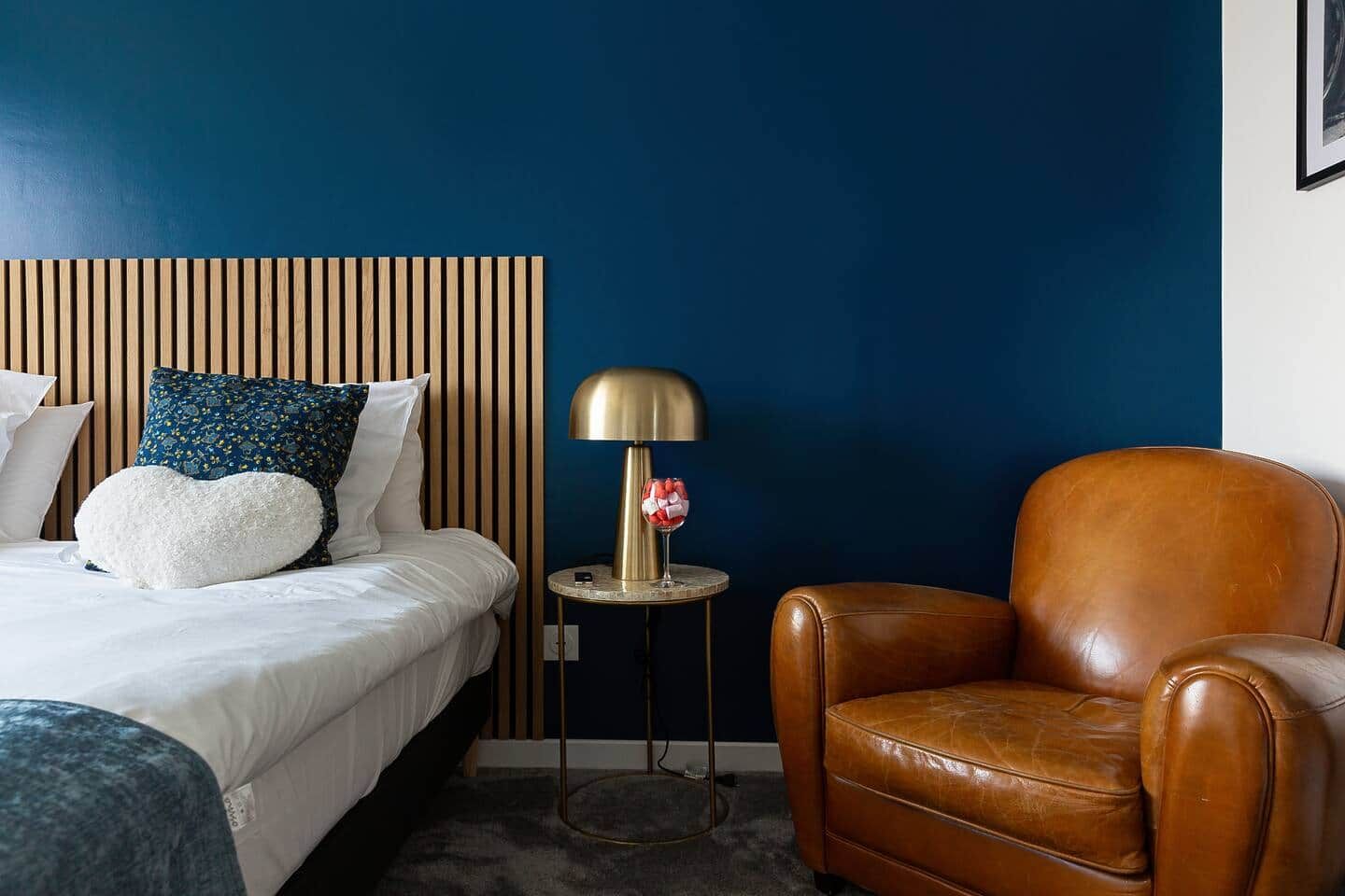 A hotel room with a bed, a brown leather armchair, a small round side table with a lamp, and a blue wall.