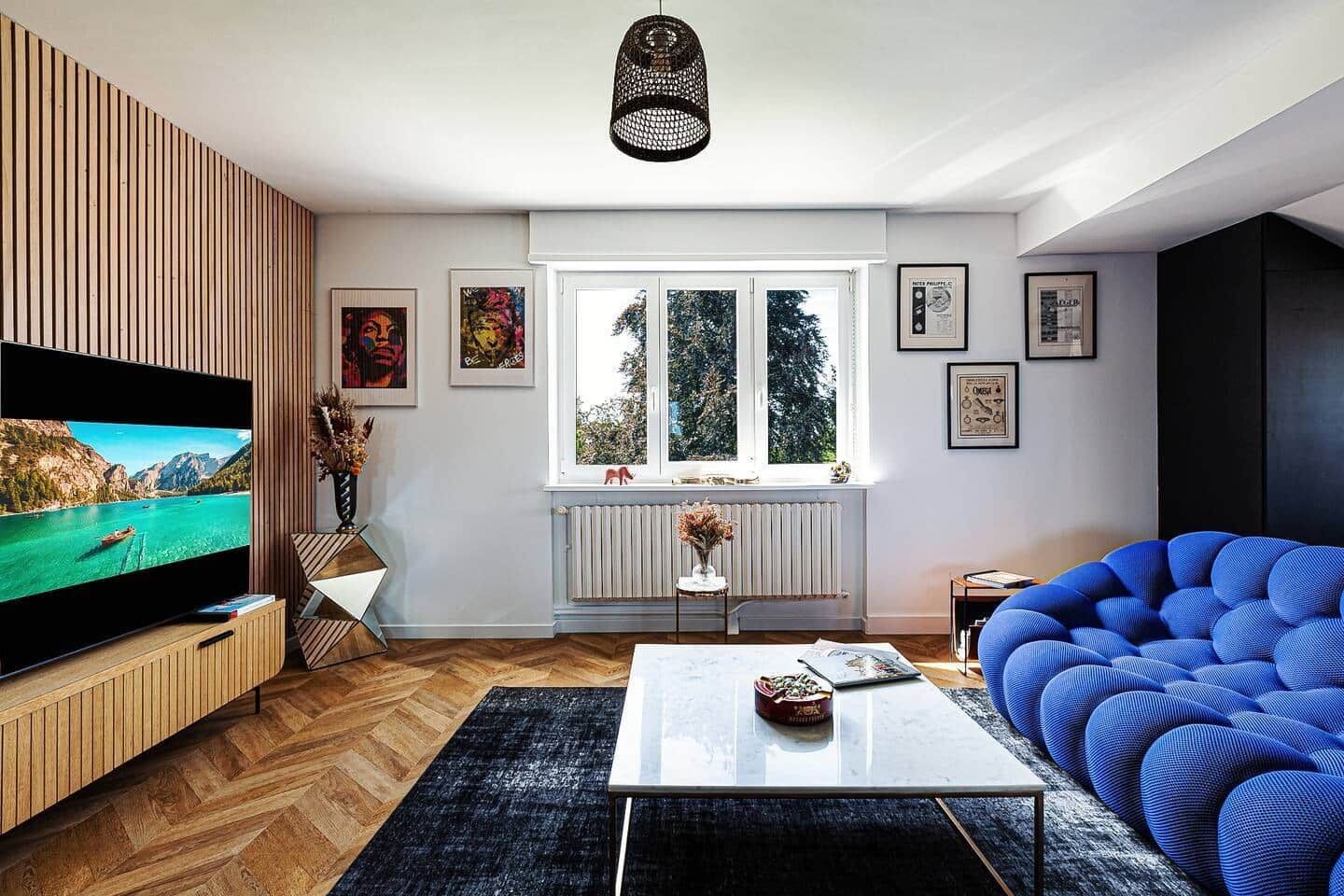 Sala de estar moderna com uma televisão de tela grande, um sofá azul, uma mesa de centro e uma janela com vista para árvores.