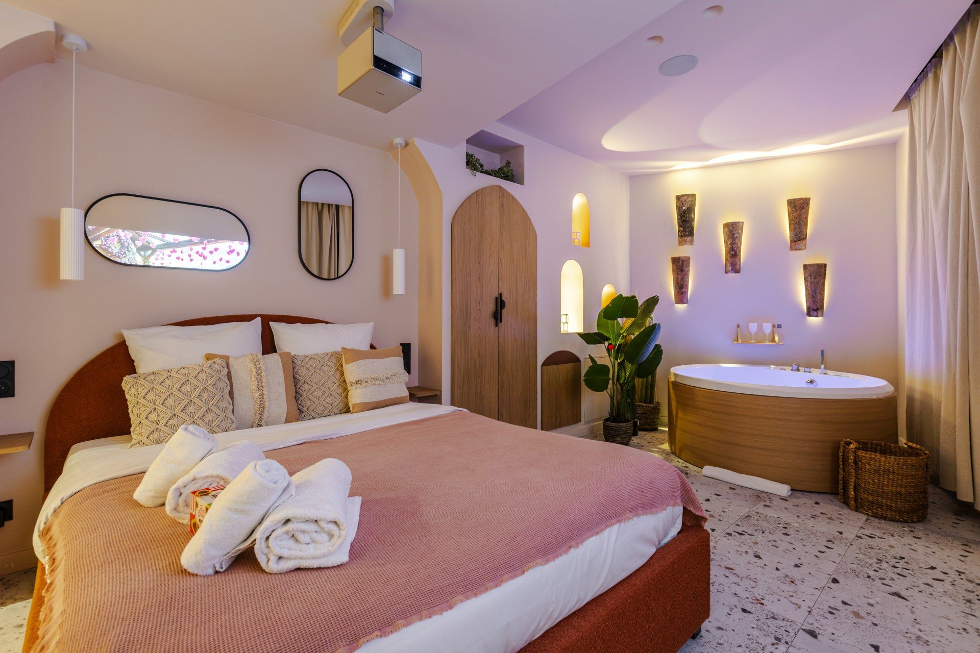 A hotel room with a large bed, a wooden wardrobe, a round mirror, a wall-mounted sink, and a potted plant.