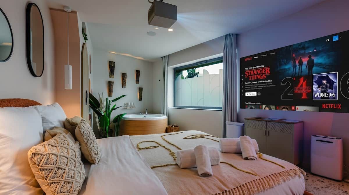 Habitació d'hotel moderna amb un llit gran, una banyera rodona, un televisor que mostra la interfície de Netflix i una finestra amb cortines.