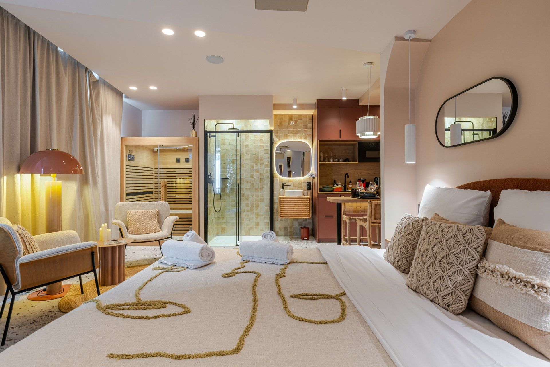 A modern hotel room with a large bed, a sitting area with a chair and a table, a bathroom with a shower and a mirror, and a kitchenette.