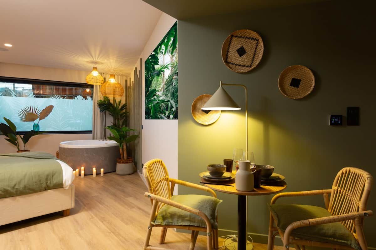 A modern hotel room with a bed, two wicker chairs, a round table, and a wall-mounted lamp.
