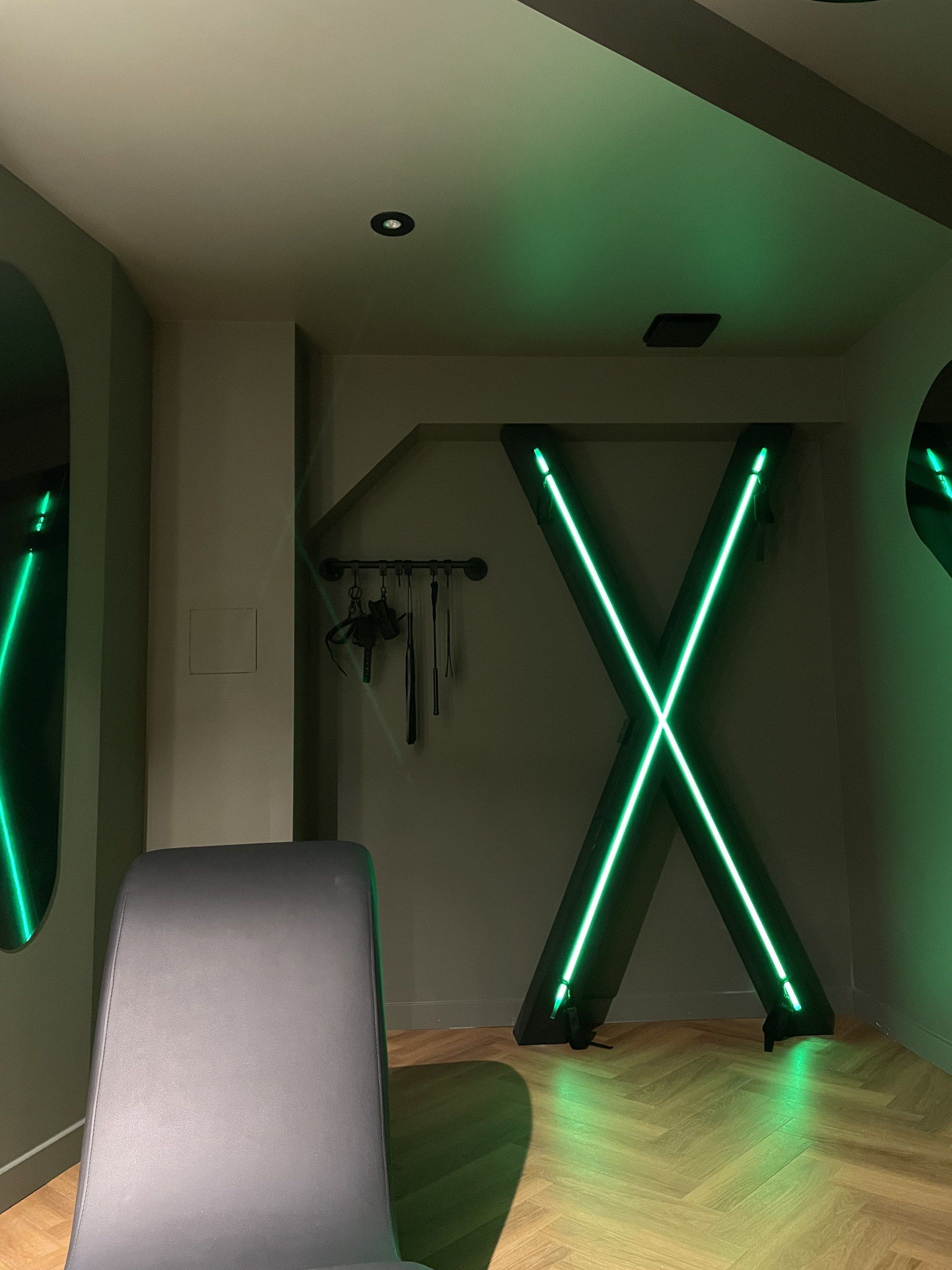 A room with a modern design, featuring a large X-shaped light installation on the wall and a grey chair in the foreground.