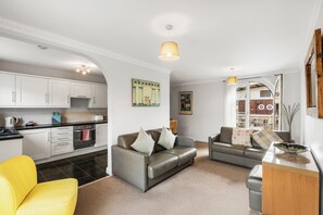 Ein modernes Wohnzimmer mit einer Couch, einem gelben Sessel, einem Essbereich mit Tisch und einer Küche mit weißen Schränken.