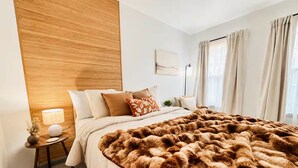 Una habitación de hotel con una cama grande, cabecera de madera, mesita de noche con lámpara y vista a la ciudad por la ventana.
