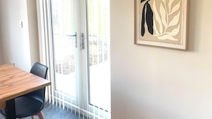 Ein Zimmer mit Holztisch, zwei Stühlen, einer Schiebeglastür und einem gerahmten Kunstwerk an der Wand.