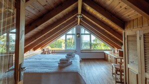 Een kamer met schuine wanden, houten lambrisering, een bed met een witte dekbedovertrek, een raam met uitzicht op groen en een houten tafel.