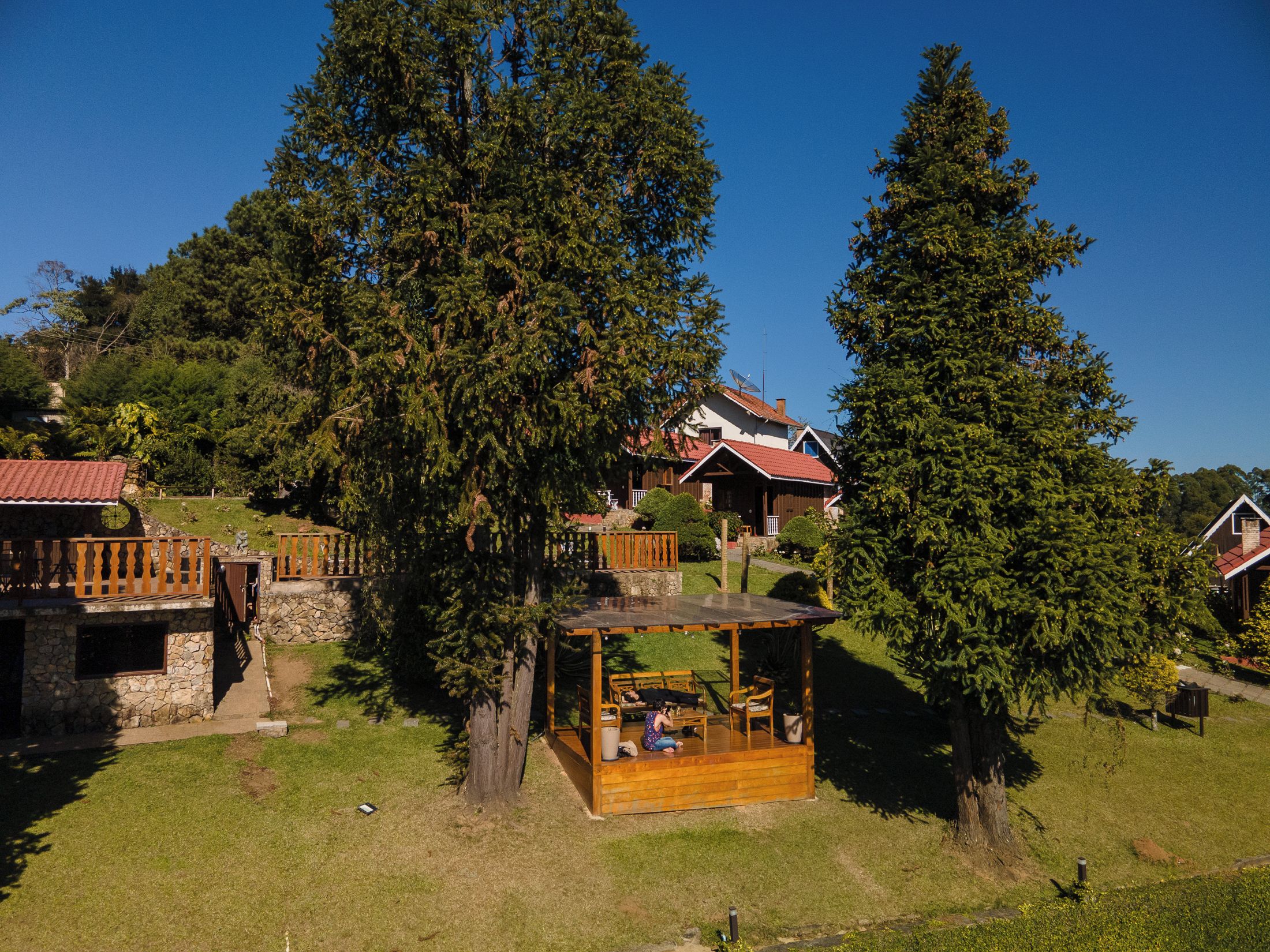 Um gazebo de madeira com bancos, cercado por árvores e um muro de pedra.