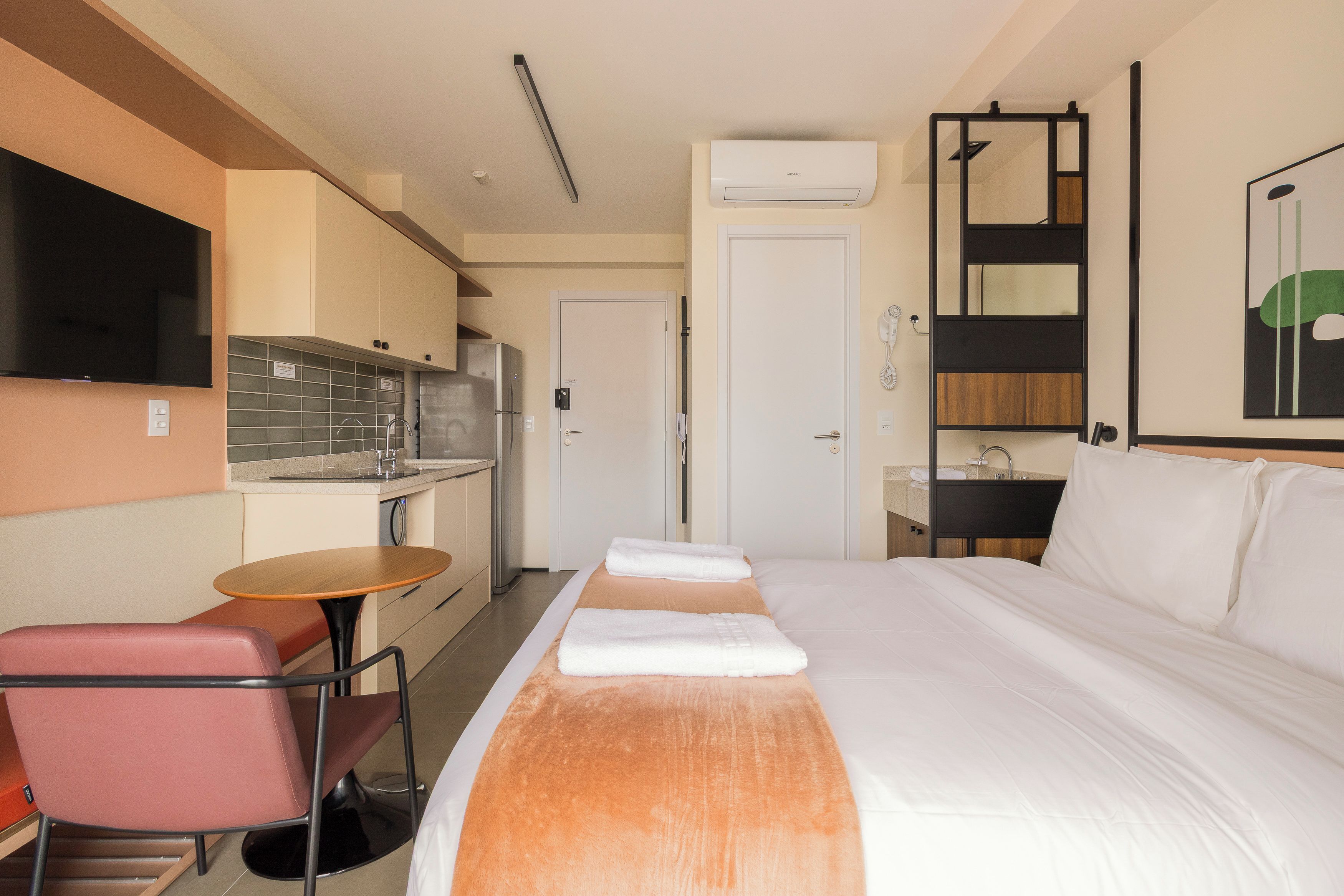 A hotel room with a bed, a small table, a chair, a TV, a kitchenette, and a bathroom.