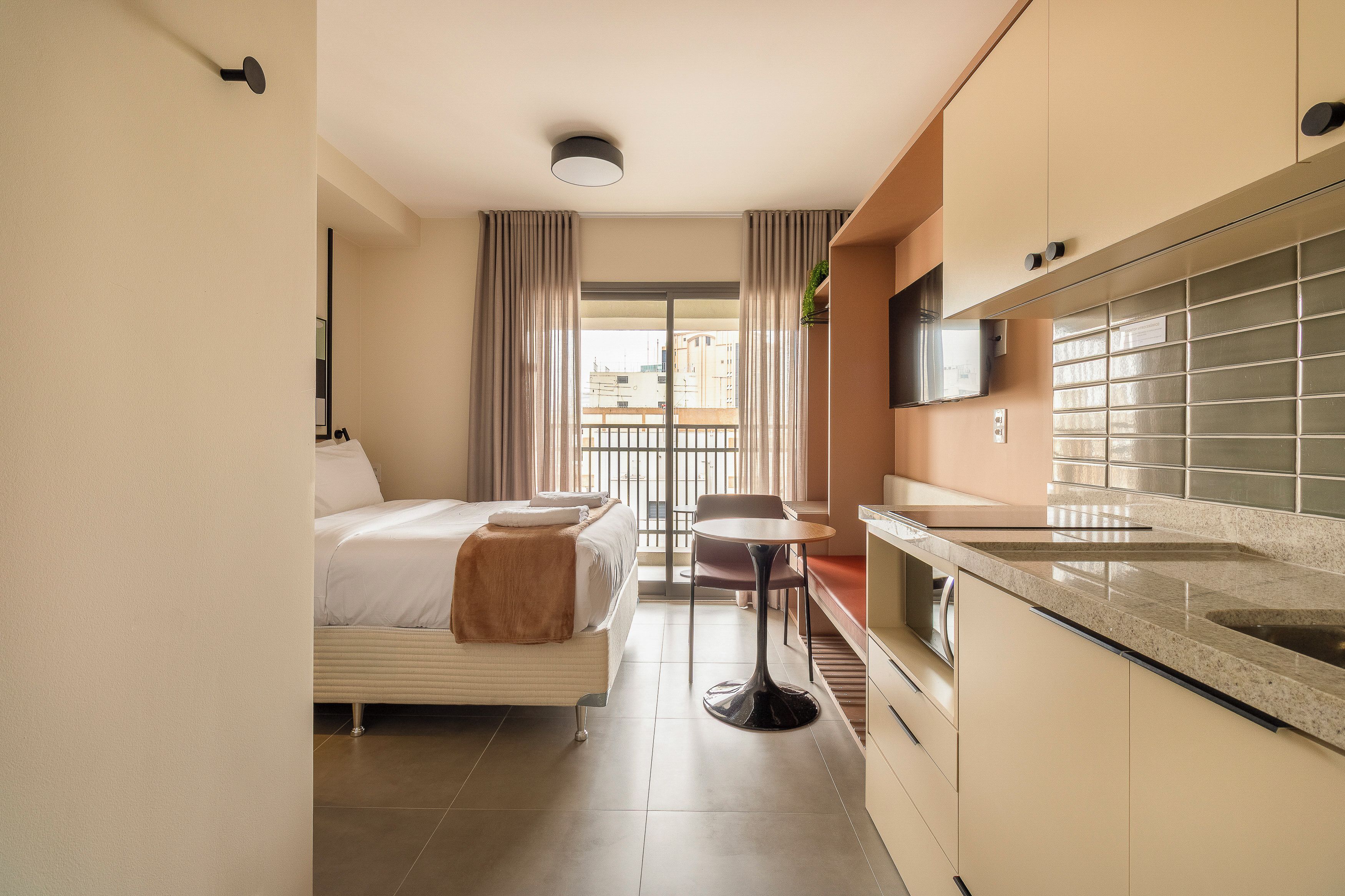 A compact hotel room with a bed, a small table, a chair, a kitchenette, and a window with curtains.