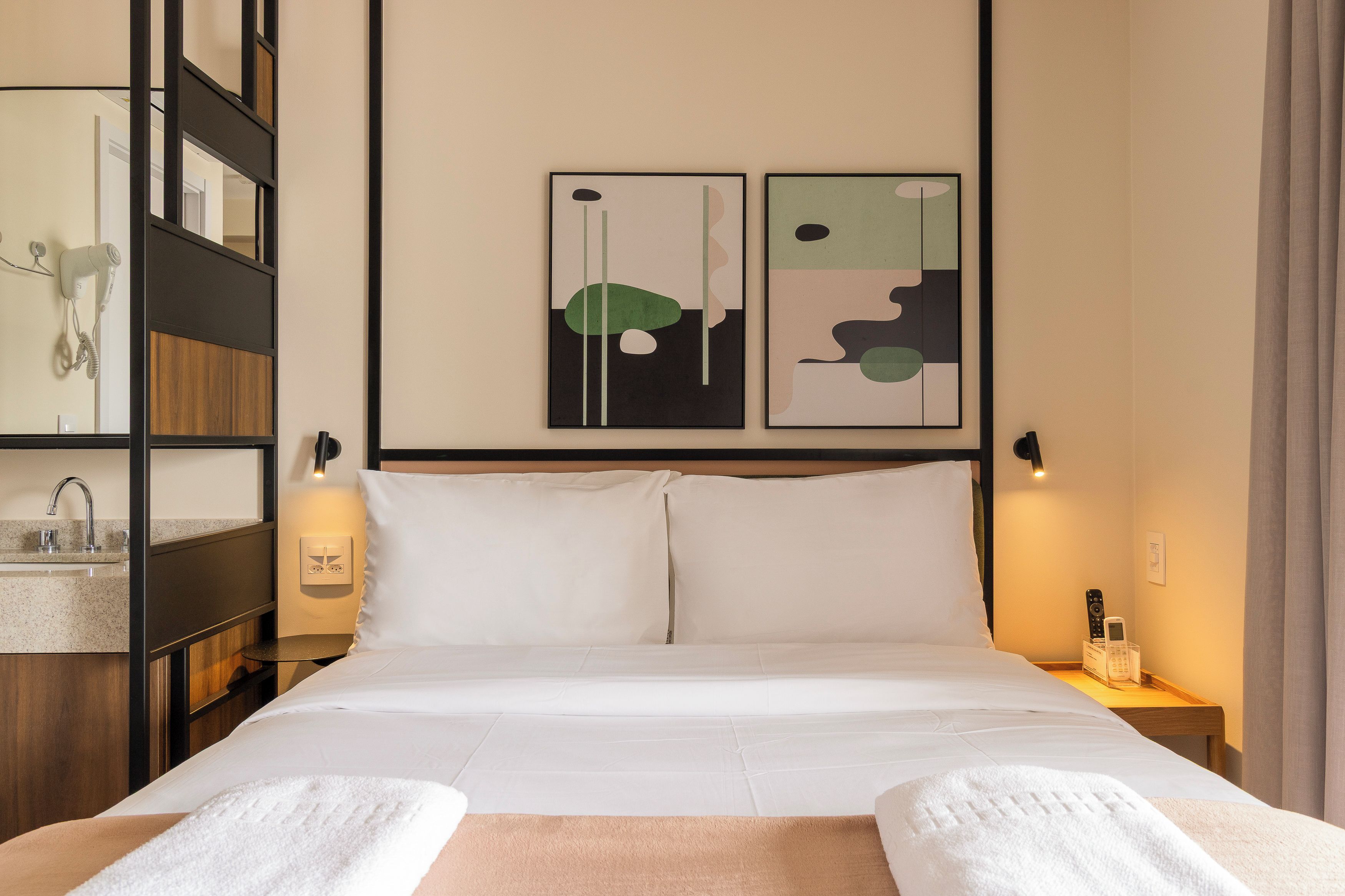 A hotel room with a bed, two wall-mounted paintings, a bedside table with a lamp, a hairdryer, and a telephone.