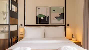 Quarto de hotel com cama, duas pinturas penduradas na parede, mesa de cabeceira com abajur, secador de cabelo e telefone.