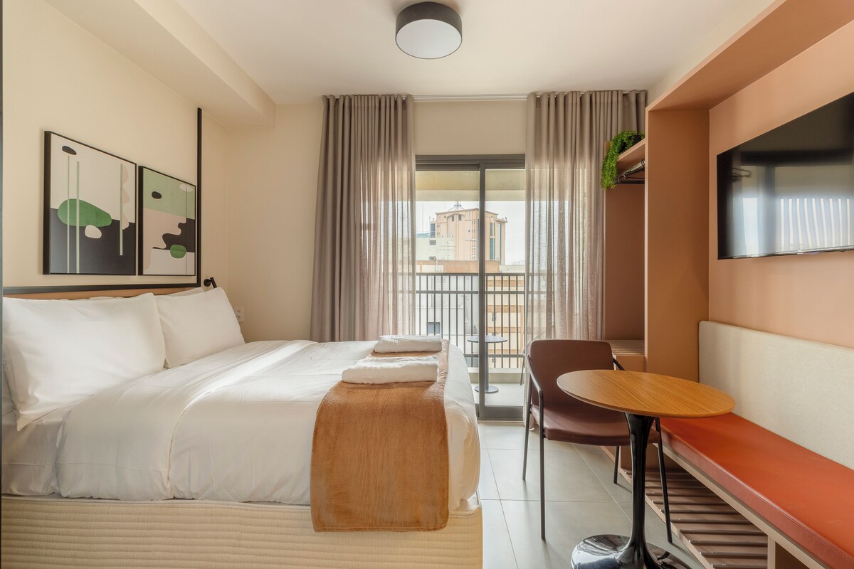 A hotel room with a bed, a chair, a small table, a TV, and a balcony with curtains.