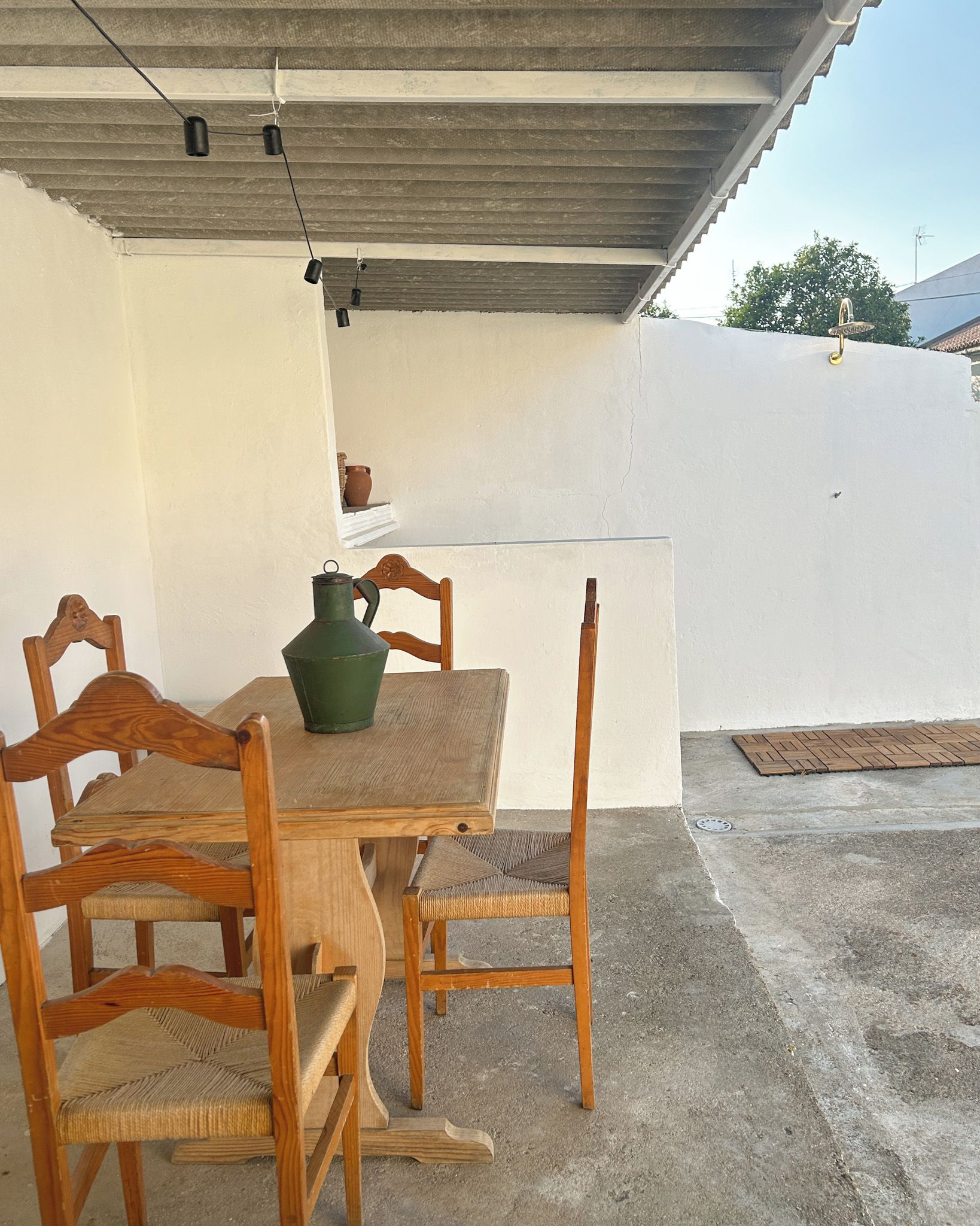 A wooden table with chairs under a corrugated metal roof, a green ceramic jug, and a window with a view of a white wall and trees.
