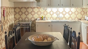 A dining area with a table set for four, a bowl of pumpkins, and a kitchen in the background with white cabinets and a patterned backsplash. - Vrbo Property (Benavila)