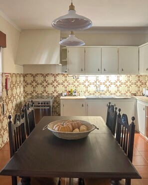 Un coin repas avec une table dressée pour quatre personnes, un saladier rempli de petites courges et, au fond, une cuisine avec des meubles blancs et un crédence à motifs.