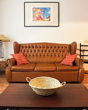 A brown leather sofa with red striped pillows, a woven basket on a wooden table, and a framed artwork above.