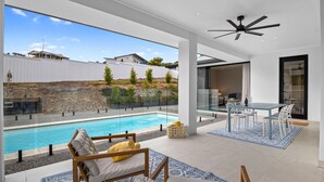 Et moderne poolområde udendørs med glasafskærmning, et spiseområde med bord og stole samt en ventilator i loftet.