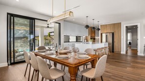 A modern kitchen with a central island, wooden flooring, and a dining area with a large table and chairs. - Jewel of Carrickalinga - 14 Dorset Court (Carrickalinga)