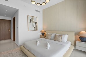 A bedroom with a large bed, two bedside lamps, a framed abstract painting, and a wooden door. - Iconic 3 Bedroom with Panoramic View in Grand Bleu (Emaar Beachfront)