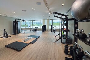 A modern gym with various exercise equipment, a wooden floor, and large windows offering a view of the outdoors. - Iconic 3 Bedroom with Panoramic View in Grand Bleu (Emaar Beachfront)